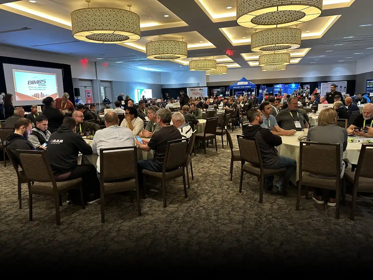 Teaching at BIMRS Conference., Conference room with attendees seated at round tables, large screens displaying a logo, and exhibition booths in the background.